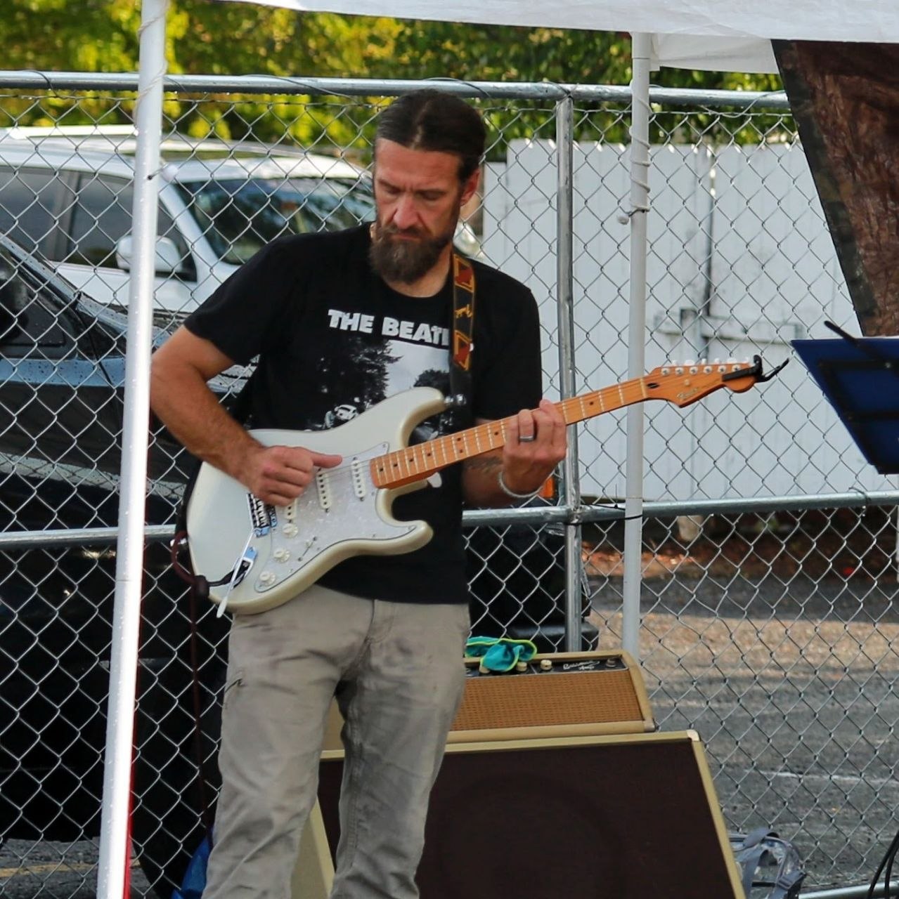 Billy Thorpe playing white Fender Stratocaster on an outdoor stage, eyes down on his hands mid-riff
